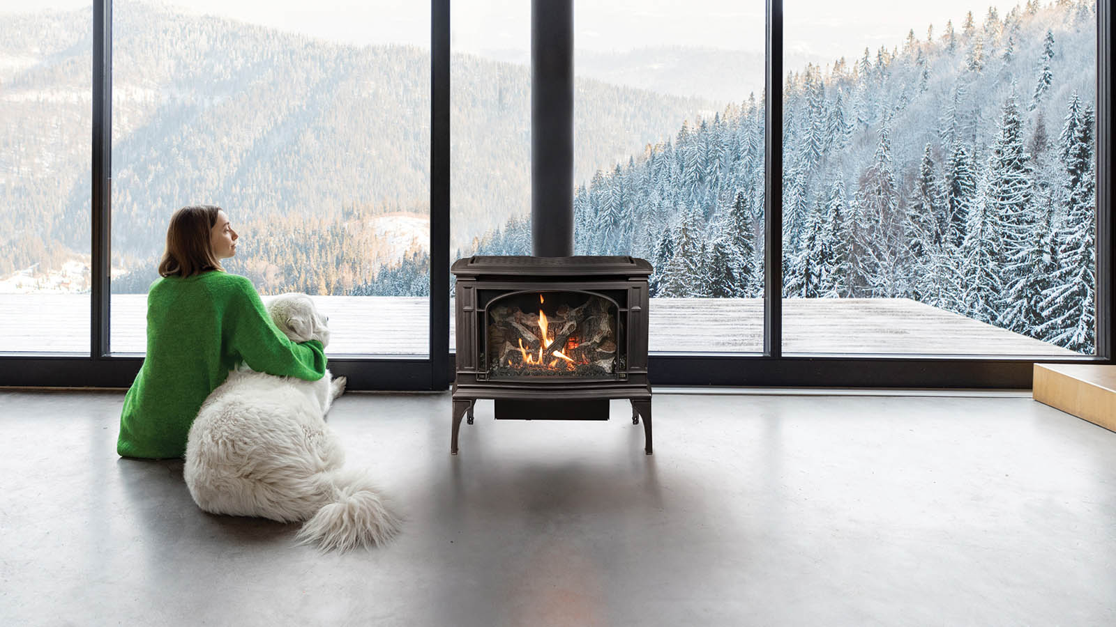 Woman in a green sweater sits with a fluffy white dog by a wood stove, gazing at a snow-covered forest through large windows.